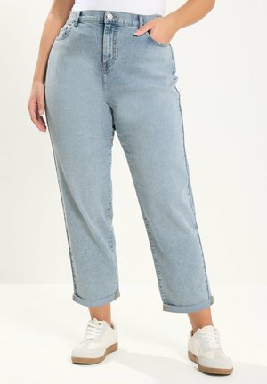 Kvinde iført lyseblå denimjeans med opbukket kant og hvide sneakers, med den ene hånd i lommen, stående på en hvid baggrund.