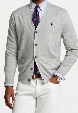 Man draagt een lichtgrijze gebreide cardigan met knopen over een blauw gestreept overhemd en een marineblauwe stropdas met rode strepen, gecombineerd met een witte broek en een polshorloge.