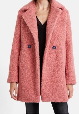 Kobieta ubrana w różowy płaszcz z wełny o średniej długości, z teksturą, szerokimi klapami i dwoma widocznymi ciemnymi guzikami, noszona na czarnej bluzce.