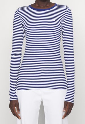 Kvinde iført en marineblå og hvid langærmet trøje med vandrette striber og hvide bukser mod en ensfarvet baggrund.