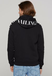 Sudadera negra con capucha y cordón, que presenta un gran texto blanco "SAILING" en la parte posterior. Puños y dobladillo acanalados, llevada con jeans de mezclilla azul.