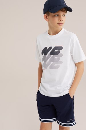 Chłopiec noszący granatową czapkę, białą koszulkę z czarnym i szarym napisem oraz granatowe spodenki z białymi paskami na krawędziach i małym detalem tekstowym.