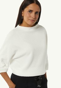 Witte, getextureerde trui met hoge hals en driekwartmouwen, gecombineerd met zwarte broek met trekkoord en metalen accenten.