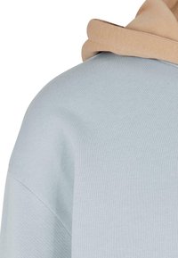 Épaule de sweat-shirt bleu clair avec capuche beige reposant dessus, montrant la texture du tissu et les détails de la couture.