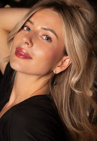 Zwarte oorbellen hangen aan het oor van het model; haar golvende, lichtblonde haar omlijst haar gezicht, dat een zachte, natuurlijke make-up look heeft.