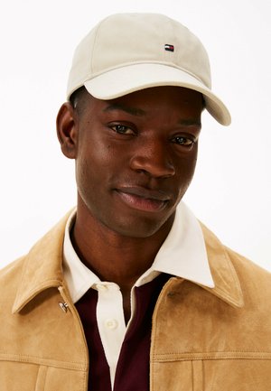 Beige Baumwollkappe mit gebogenem Schirm, die ein kleines Logo-Patch trägt, getragen mit einer braunen Wildlederjacke über einem weiß-rot karierten Hemd.