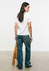 T-shirt blanc à manches courtes, jeans évasés en denim bleu, cardigan marron texturé porté sur le bras et chaussures à motifs. Design simple et épuré.