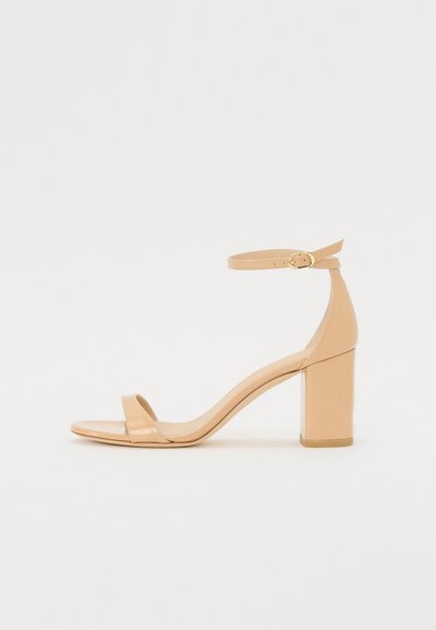 Sandales à talons en cuir verni nude avec un orteil carré, une sangle à la cheville et un talon bloc chunky. Texture lisse et détails de design minimalistes.