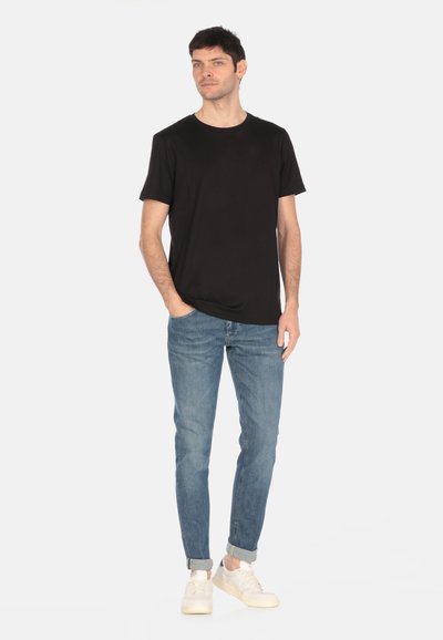 T-shirt nera a girocollo, maniche corte, abbinata a jeans in denim azzurro chiaro e sneakers bianche, in piedi su uno sfondo semplice.