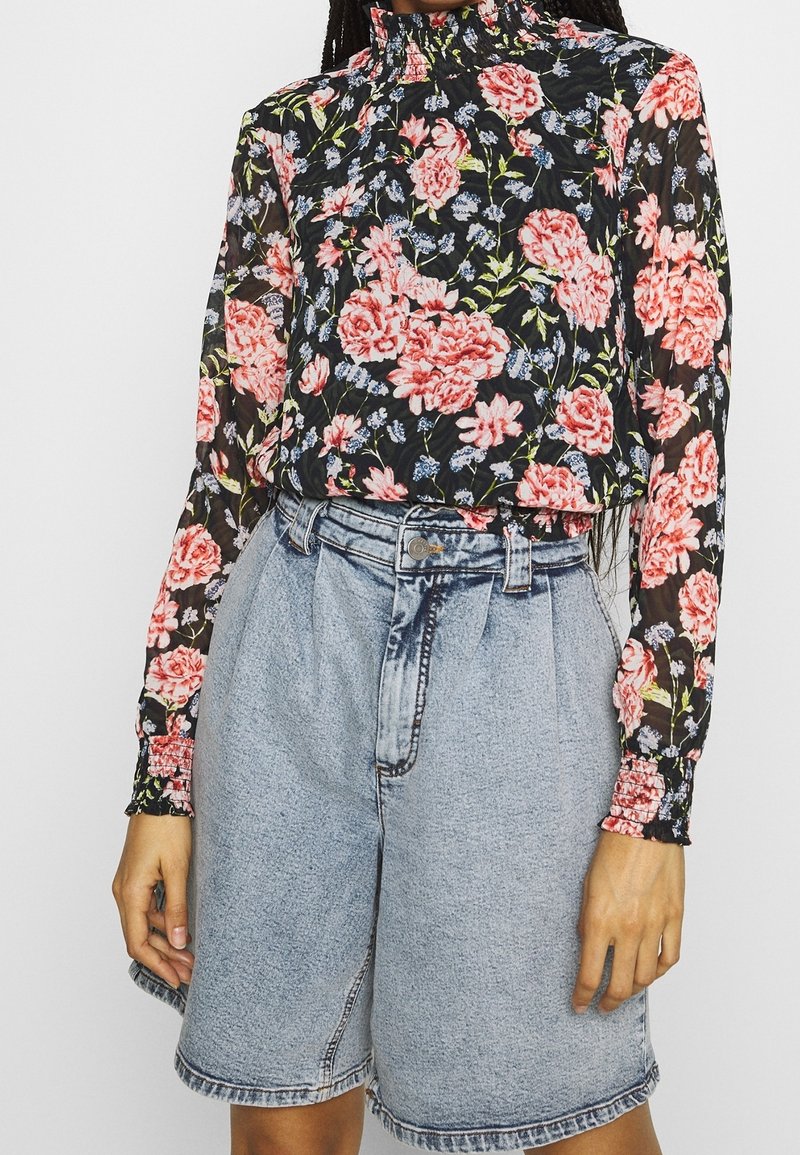 Bluse mit langen Ärmeln und floralen Motiven in Schwarz, mit einem hohen Kragen, verziert mit rosa und blauen Blumen. Kombiniert mit hellblauen, locker sitzenden Denim-Shorts.