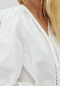 Blusa bianca in cotone con scollo a V, chiusura con bottoni e dettagli in pizzo intricati lungo le cuciture. Le maniche a sbuffo aggiungono volume.