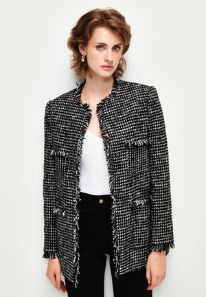 Twee blazer in zwart-witte houndstoothprint, met rafelige randen, open voorkant en twee opgestikte zakken. Draag het over een witte top en zwarte jeans.