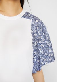 Camiseta blanca con mangas abullonadas con un patrón floral azul, con cuello redondo y textura suave. Las mangas tienen detalles recogidos para dar volumen.