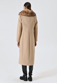 Cappotto beige con colletto in pelliccia sintetica, design dritto e maniche lunghe. Indossato con stivali neri alti fino al ginocchio con un accento a catena.