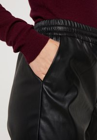 Zwarte nep leren broek met een elastische tailleband, voorzien van een zijzak en een gladde textuur. Bordeaux kleurige gebreide top met lange mouwen zichtbaar.