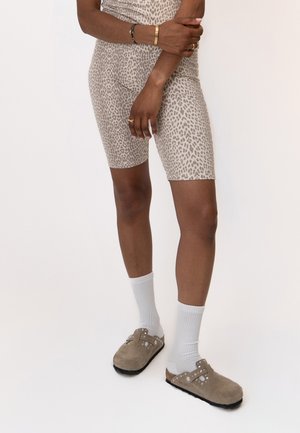 Personne portant un short et un haut beige à motif léopard, avec des chaussettes blanches et des chaussures beiges sans lacets, debout les bras croisés.