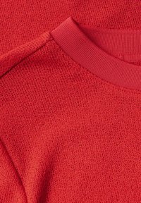 Červená pletená tkanina s texturovaným povrchem; obsahuje hladký žebrovaný límec ve shodné barvě, což vytváří jednotný design.