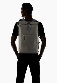 Zaino roll-top in tessuto grigio chiaro, con una tasca frontale con cerniera e tirante rosso, spallacci regolabili e un design minimalista.