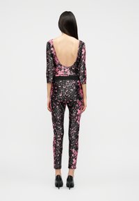 Γυναίκα που στέκεται, φοράει ολόσωμη φόρμα με ροζ και μαύρο μοτίβο, με βαθύ ανοικτό πίσω μέρος και μαύρες ψηλοτάκουνες γόβες, γυρισμένη πλάτη.