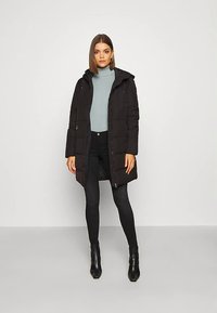 Donna che indossa un lungo cappotto trapuntato nero con cappuccio, dolcevita blu chiaro, pantaloni skinny neri e stivaletti neri, in piedi di fronte a una parete bianca.