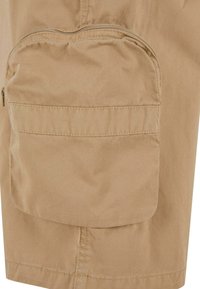 Shorts cargo beiges avec une grande poche arrondie zippée sur la cuisse, fabriqués en tissu léger. Présente des coutures visibles et une coupe décontractée.