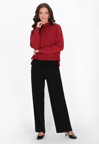 Pulover roșu tricotat cu guler înalt și accesorii la decolteu, purtat cu pantaloni negri evazați și tocuri negre.