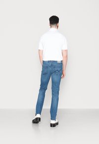 Man står vänd bort, iförd vit kortärmad skjorta, blå slim-fit jeans och vita platåstövlar mot en enfärgad bakgrund.