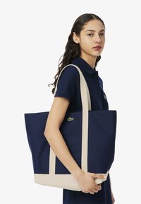 Sac en toile bleu marine avec des poignées et une base beiges, arborant un petit logo vert sur le devant. Forme rectangulaire avec le haut ouvert.