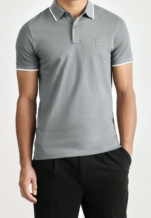 Mężczyzna w szarej koszulce polo z białym wykończeniem na kołnierzyku i rękawach, w czarnych spodniach, z rękami swobodnie opuszczonymi wzdłuż ciała.