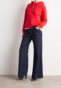 Czerwona bawełniana bluza z kangurzą kieszenią, zestawiona z ciemnoniebieskimi spodniami o szerokich nogawkach. Czarne szpilki o spiczastym nosku dopełniają całość.
