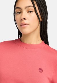 Sudadera rosa con cuello redondo y puños de canalé. Presenta un logo bordado en el pecho. Textura suave y diseño casual.