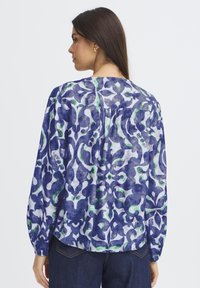 Femme aux longs cheveux bruns portant un chemisier à manches longues bleu, blanc et vert à motifs et un jean bleu foncé, tournée de côté devant un fond blanc.