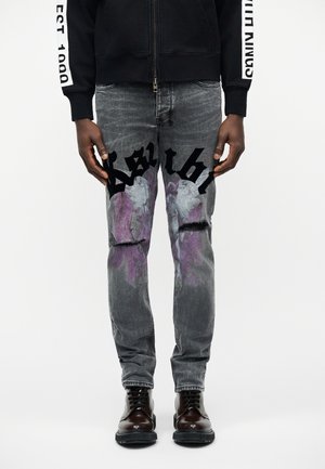 Homme portant un jean gris usé avec des textes gothiques noirs et des motifs d'ailes d'ange violettes, des bottes brun foncé et une veste zippée noire.