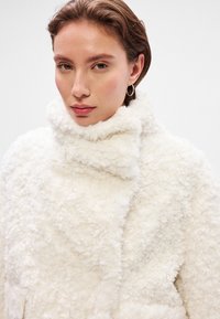 Λευκή, αφράτη παλτό με ψηλό, κυκλικό γιακά. Η υφή είναι απαλή και γουνωτή, με ασυμμετρικό κλείσιμο μπροστά και χωρίς ορατό εξοπλισμό.