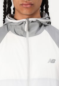Jachetă sport albă cu Accente gri, ușoară, cu fermoar în față, design cu glugă, are logo-ul New Balance pe partea stângă a pieptului.