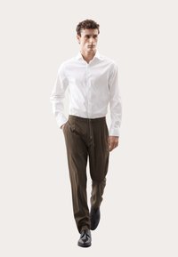 Weißes Hemd mit Knopfleiste, kombiniert mit olivgrünen Jogginghosen mit Kordelzug. Das Model trägt schwarze Anzugschuhe. Einfaches Design, glatte Textur und lässige Passform.