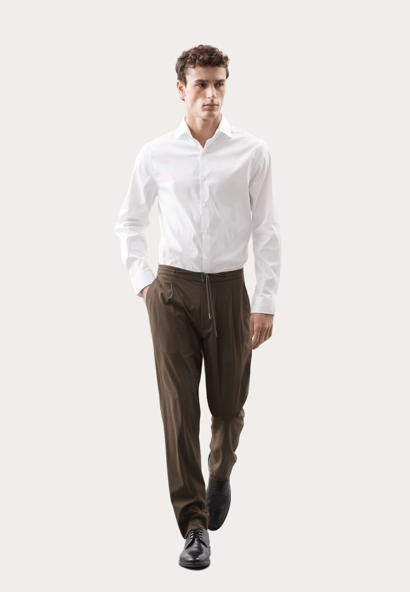 Weißes Hemd mit Knopfleiste, kombiniert mit olivgrünen Jogginghosen mit Kordelzug. Das Model trägt schwarze Anzugschuhe. Einfaches Design, glatte Textur und lässige Passform.