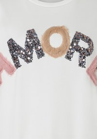 Weißer Stoff mit dem Wort "MOM", wobei das M und das P aus schwarzen Pailletten bestehen und das O aus beigem, flauschigem Material sowie rosa, flauschigen Kanten gefertigt ist.