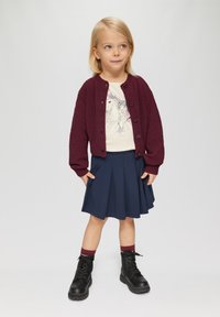 Bordeaux gebreide cardigan, crèmekleurig shirt met grafische print, marineblauwe geplooide rok en zwarte laarzen met rode sokaccenten.
