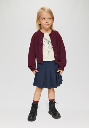 Bordeaux gebreide cardigan, crèmekleurig shirt met grafische print, marineblauwe geplooide rok en zwarte laarzen met rode sokaccenten.