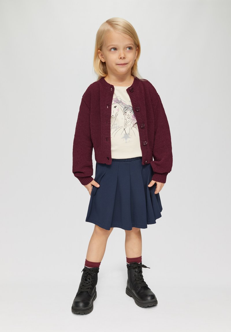 Bordeaux gebreide cardigan, crèmekleurig shirt met grafische print, marineblauwe geplooide rok en zwarte laarzen met rode sokaccenten.