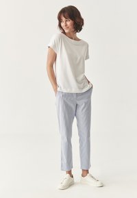 White short-sleeve t-shirt paired with light blue striped trousers. Model wears white sneakers, hands in pockets, with a relaxed stance.