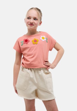 Jeune fille debout, main sur la hanche, portant un t-shirt orné de fleurs roses et un short beige avec poches avant, sur fond blanc.