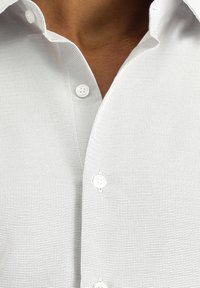 Camisa blanca de texturizado con un pequeño patrón de cuadros, con botones blancos y un diseño de cuello clásico.