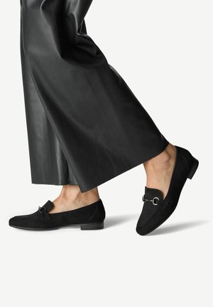 Mocassins en daim noir à talon bas, avec un accent décoratif en métal sur le dessus. Portés avec un pantalon large en cuir noir.