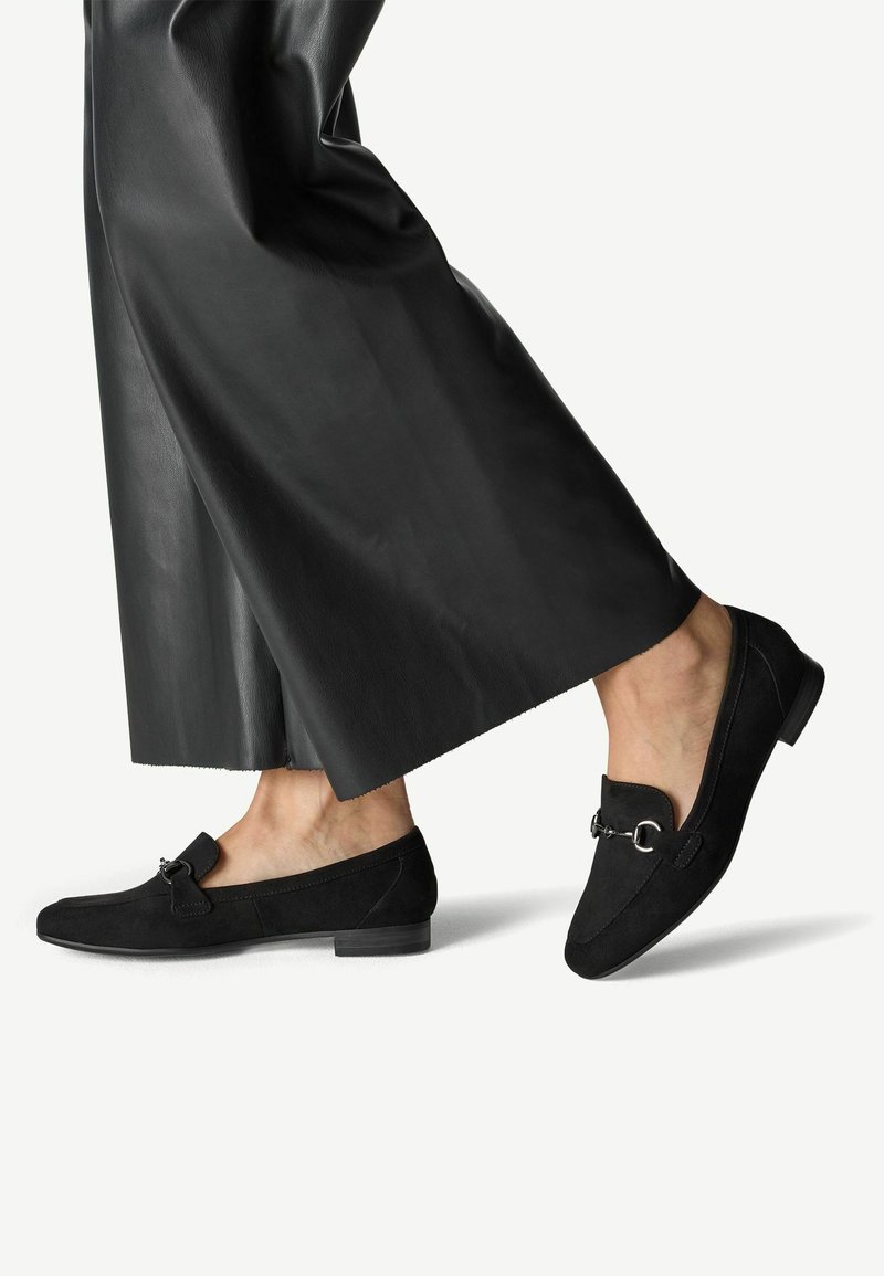 Mocasines de ante negro con un tacón bajo, que cuentan con un acento de hardware decorativo en la parte superior. Se llevan con pantalones anchos de cuero negros.