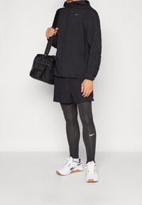 Chaqueta ligera negra con capucha, combinada con pantalones cortos negros y mallas de compresión negras. Zapatillas deportivas blancas con suela marrón. Bolsa de gimnasio negra.
