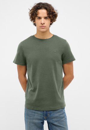 Dunkelgrünes T-Shirt mit kurzen Ärmeln und rundem Halsausschnitt, aus weichem Stoff, mit einem kleinen Logo auf der linken Brust. Getragen mit blauen Jeans.