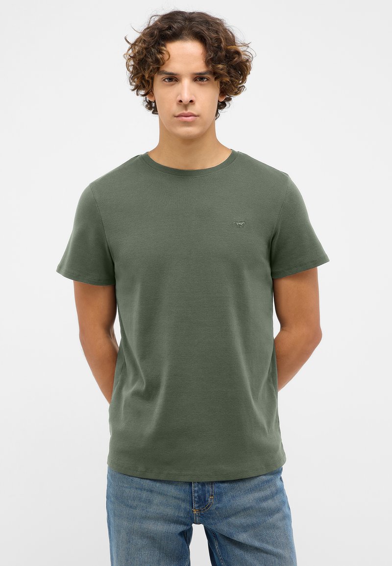 T-shirt court à manches courtes de couleur vert foncé avec un col rond, en tissu doux, arborant un petit logo sur la poitrine gauche. Porté avec un jean bleu.