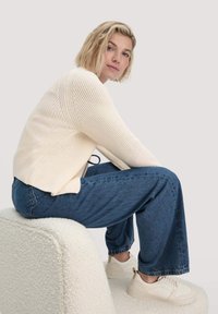 Hellbeige gestrickter Pullover mit einem strukturierten Muster, kombiniert mit blauen weit geschnittenen Jeans und beigen Sneakers, sitzt auf einem weichen, strukturierten Hocker.